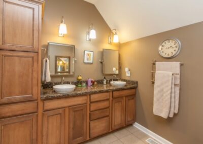 A well-lit bathroom with dual sinks, wooden cabinets, and a large wall clock.