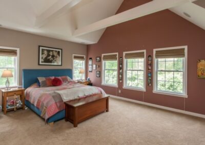 A welcoming bedroom featuring a blue upholstered bed, terracotta walls, multiple windows, and exposed ceiling beams.