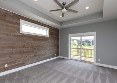 Empty modern room with a wooden accent wall, gray carpeting, and a ceiling fan, leading to a balcony with a neighborhood view.