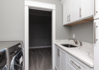 A modern laundry room with white cabinetry and appliances, featuring a sink and an open door leading to a dark room.